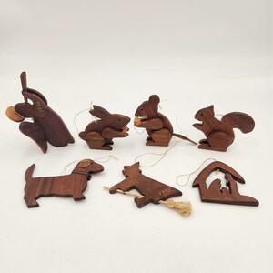 May include: A set of seven wooden Christmas ornaments featuring various animals, including a rabbit, squirrel, mouse, dog, and a witch on a broomstick. The ornaments are all hand-carved and painted in a dark brown color. The ornaments are dated 1975 and 1981.