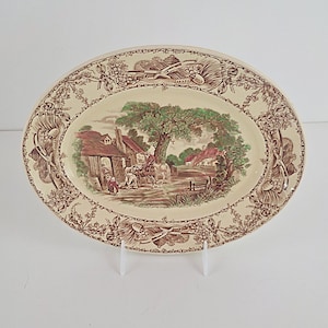May include: Oval serving platter with a pastoral scene in brown and green tones. The center features a village scene with a large tree, houses, and figures. The rim has a decorative floral and scroll design.