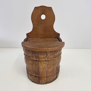 May include: A vintage wooden salt box. The box has a curved back with a round hole at the top. The barrel-shaped body is made of vertical and horizontal wooden slats. The wood has a warm, natural tone.