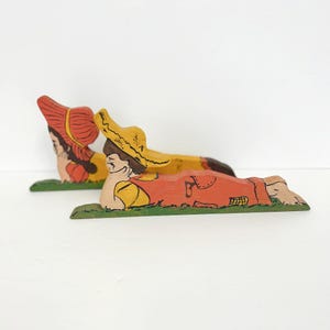 May include: Two wooden figurines of children lying on their sides, painted in bright colours. The girl is wearing a red dress and a yellow hat, while the boy is wearing a yellow shirt and red overalls. Both figurines are lying on a green painted base.