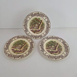 May include: Three antique ceramic plates with a cream-colored background and a brown forest scene. The plates have a decorative brown floral border. The plates are round and oval shaped.