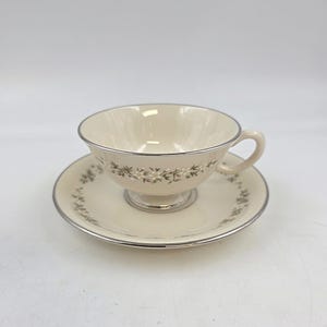 May include: Cream-colored teacup and saucer set with a silver trim. The cup and saucer feature a delicate floral pattern in a ring around the rim. The teacup has a small handle and sits on a pedestal base.