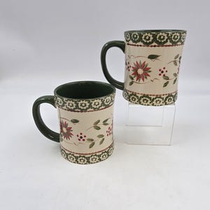 May include: Two ceramic mugs with a cream-colored base, adorned with a floral design featuring red flowers, green leaves, and red berries. The rims and handles are dark green. The mugs are displayed on a clear stand against a white background.