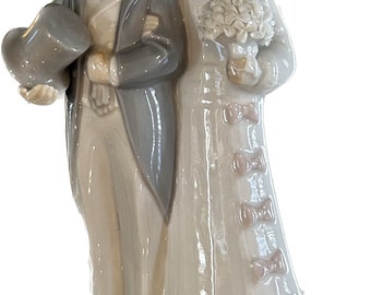 May include: A porcelain figurine of a bride and groom in traditional wedding attire. The bride is wearing a white dress with a lace trim and a veil. The groom is wearing a black suit with a white shirt and a bow tie. They are holding hands and smiling.