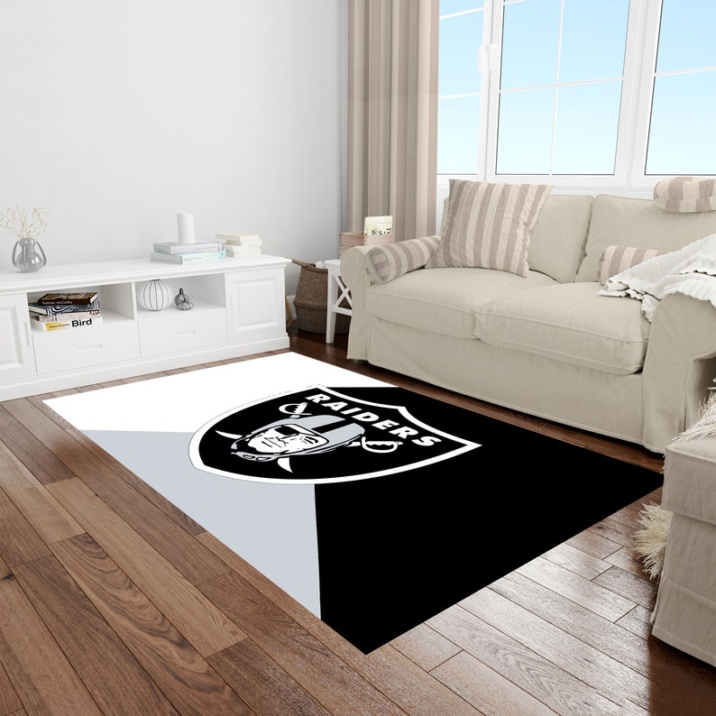 May include: Black and white area rug with the Las Vegas Raiders logo. The logo features a silver helmet with a skull and crossbones, and the word "RAIDERS" written in a white arc above the helmet.