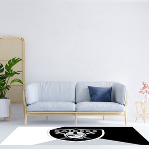 May include: A black and white area rug with the Las Vegas Raiders logo in the center. The rug is on a white floor in front of a light blue sofa with a blue pillow.