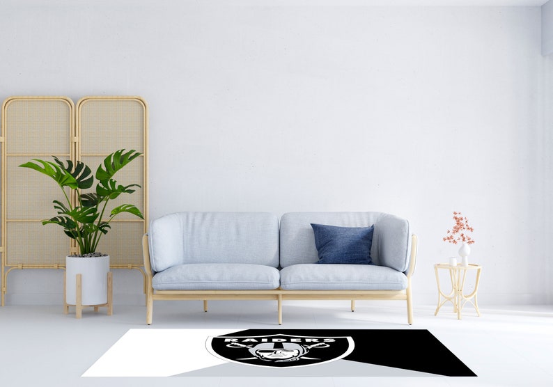 May include: A black and white area rug with the Las Vegas Raiders logo in the center. The rug is on a white floor in front of a light blue sofa with a blue pillow.