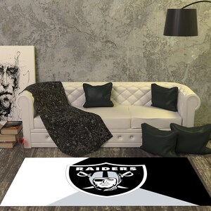 May include: A black and white rug with the Raiders logo in the center. The logo is a silver shield with two crossed swords and a skull with a helmet on top. The text "RAIDERS" is above the shield. The rug is on a wooden floor in a living room setting.