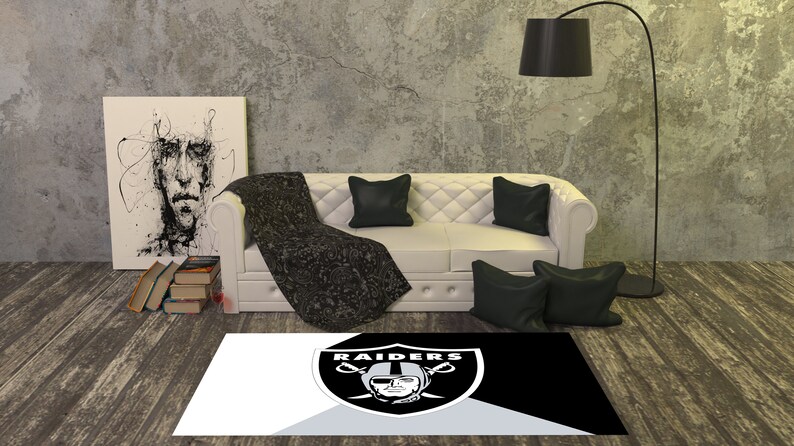 May include: A black and white rug with the Raiders logo in the center. The logo is a silver shield with two crossed swords and a skull with a helmet on top. The text "RAIDERS" is above the shield. The rug is on a wooden floor in a living room setting.