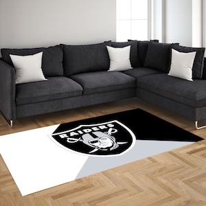 May include: A black and white area rug with the Las Vegas Raiders logo in the center. The logo is a silver shield with the word "RAIDERS" written across the top and a player's face in the center. The rug is on a hardwood floor in front of a gray sectional sofa.
