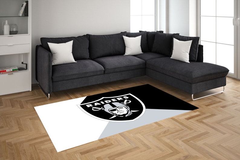May include: A black and white area rug with the Las Vegas Raiders logo in the center. The logo is a silver shield with the word "RAIDERS" written across the top and a player's face in the center. The rug is on a hardwood floor in front of a gray sectional sofa.