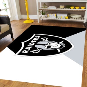 May include: Black and white rug with the Raiders logo and a player's face in the center. The logo is on a black shield with the word "RAIDERS" written in white.