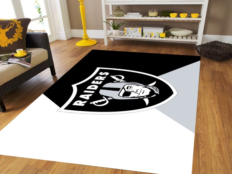 May include: Black and white rug with the Raiders logo and a player's face in the center. The logo is on a black shield with the word "RAIDERS" written in white.