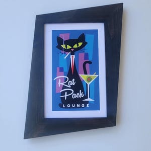 May include: A framed art print featuring a stylized black cat smoking a cigarette and wearing a red tie, with a martini glass. The print has the words "Rat Pack Lounge" and is set against a blue and pink background.