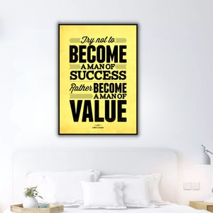 May include: A framed poster with the quote "Try not to become a man of success. Rather become a man of value" in black text on a yellow background. The poster is hanging above a white bed in a bedroom.