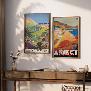 K&ouml;nnte beinhalten: Zwei Vintage-Reiseposter in Holzrahmen. Das Poster links ist eine farbenfrohe Illustration einer Bergstadt mit dem Text "Vence Hiver Ete Cote D'Azur". Das Poster rechts ist eine farbenfrohe Illustration einer Strandszene mit dem Text "Annecy La Plage".