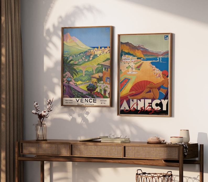 K&ouml;nnte beinhalten: Zwei Vintage-Reiseposter in Holzrahmen. Das Poster links ist eine farbenfrohe Illustration einer Bergstadt mit dem Text "Vence Hiver Ete Cote D'Azur". Das Poster rechts ist eine farbenfrohe Illustration einer Strandszene mit dem Text "Annecy La Plage".