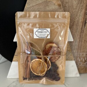 Könnte beinhalten: Ein klarer Zellophanbeutel mit getrockneten Orangenscheiben, Zimtstangen und anderen Potpourri-Zutaten, mit Bindfaden gebunden. Auf dem Beutel steht "Simmer Pot". Der Beutel wird vor einer Weihnachtsbaumdekoration aus Holz präsentiert.