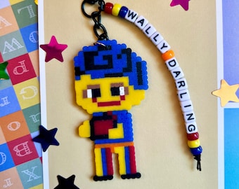 Può includere: Un portachiavi in pixel art giallo e blu con un personaggio con un cappello blu e il nome "WALLY DARLING" scritto su una catena di perline.