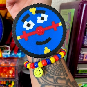May include: A blue and yellow pixel art smiley face charm on a red, yellow, and blue beaded bracelet.