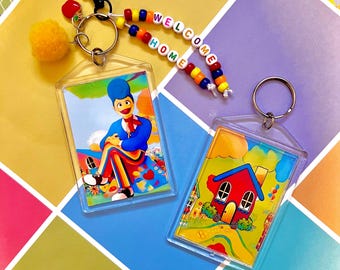 Può includere: Due portachiavi in acrilico trasparente con immagini di cartoni animati. Uno presenta un personaggio seduto in blu, rosso e colori arcobaleno. L'altro mostra una casa rossa con un tetto blu. Un filo di perline recita "WELCOME HOME". Sono attaccati anche un pompon giallo e una clip nera.