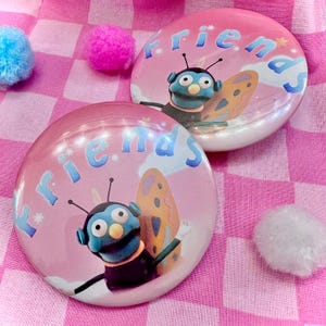 May include: Two round, pink buttons with the word "friends" in blue script. Each button features a cartoon character with blue skin, antennae, and butterfly wings. The background is a pink and white checkered pattern.