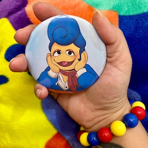 May include: A blue and white button featuring a cartoon character with blue hair, a yellow face, and a red scarf. The character is smiling and has their hands on their cheeks.
