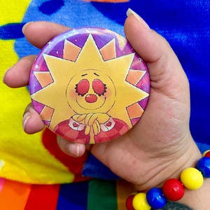 May include: A yellow sun with a red nose and red eyes wearing a red and white plaid shirt with hearts on the collar. The sun is smiling and has a pink and purple background with white stars.