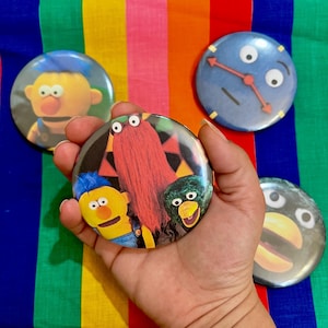 May include: Four round button pins featuring characters from the children's television show Sesame Street. The pins feature Elmo, Big Bird, Cookie Monster, and a blue character with a clock face.