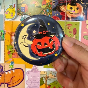 May include: A circular pinback button featuring a dark blue background with a smiling crescent moon and a bright orange jack-o'-lantern wearing a black witch hat. The design includes small yellow stars scattered across the dark blue night sky.