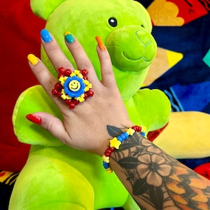 Könnte beinhalten: Eine Hand trägt einen roten, gelben und blauen Perlenring mit einem Smiley-Anhänger. Die Hand trägt auch ein blaues, gelbes und rotes Perlenarmband mit Stern-Anhängern. Die Hand ruht auf einem grünen Plüschteddybär.