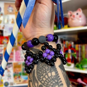 May include: Two beaded bracelets on a wrist. The top bracelet is purple and black with a black charm. The bottom bracelet is a black and purple kandi cuff with a geometric pattern.