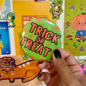 May include: A round green pinback button featuring the orange text TRICK OR TREAT and small orange star shapes scattered across the surface. The button has a glossy finish and is held against a background of various colorful illustrated stickers.