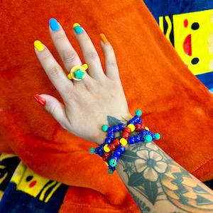 Könnte beinhalten: Ein buntes Perlenarmband mit blauen, roten, gelben und grünen Perlen. Das Armband befindet sich am Handgelenk einer Person mit einem grünen Sternring am Finger.
