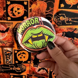 May include: A circular pin-back button featuring a graphic illustration of bright green lips with sharp yellow fangs. The word HORROR is printed in bold lime green letters along the top edge of the design, set against a dark background.