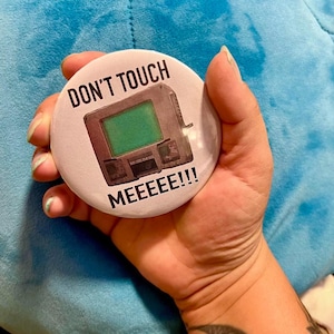 Op de afbeelding: Een witte knop met een retro computer afbeelding en de tekst "DON'T TOUCH MEEEEEEE!!!"