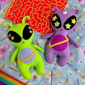 Könnte beinhalten: Zwei handgefertigte Alien-Plüschpuppen. Eine ist limettengrün, die andere lila. Beide haben große schwarze Augen mit Sternakzenten, ein Planetendesign und Antennen. Sie liegen auf einer hellblauen Oberfläche mit Blumenmuster.