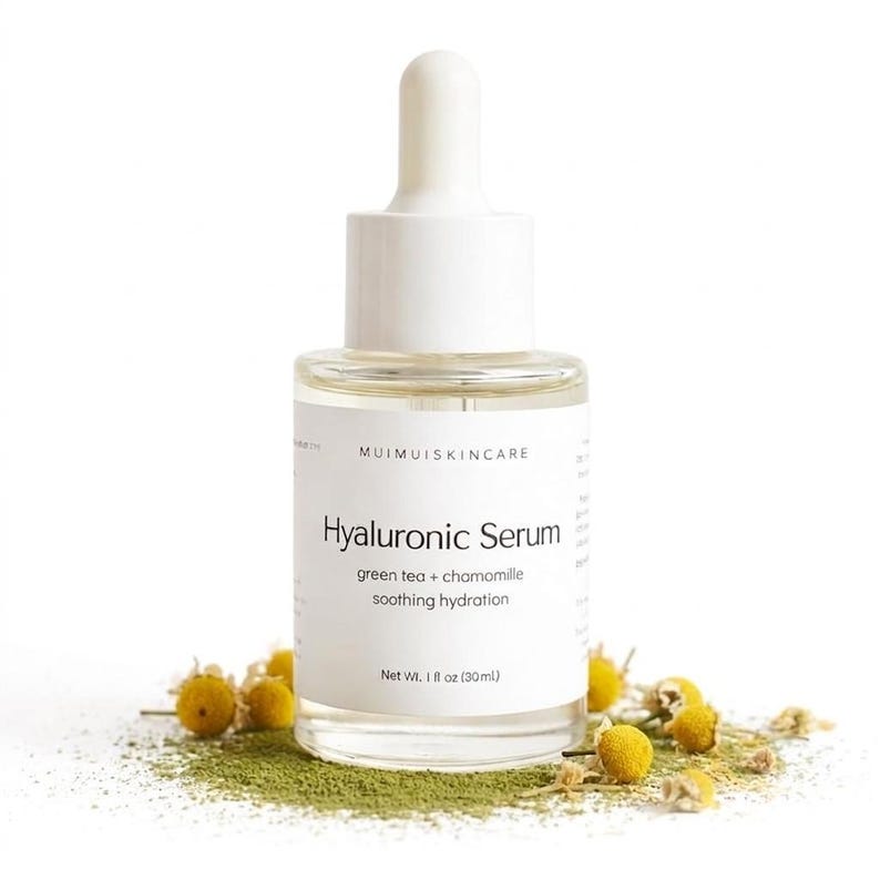 May include: A clear glass bottle of Hyaluronic Serum with a white dropper and cap. The label reads "Hyaluronic Serum" with "green tea + chamomile" and "soothing hydration" below. The bottle contains 1 fl oz (30ml). Chamomile flowers and green powder surround the bottle.