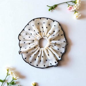 May include: A pleated hair scrunchie in off-white fabric featuring a pattern of small black polka dots. The circular accessory has a ruffled texture and a thin black stitched edge along the outer perimeter, creating a gathered, voluminous appearance.
