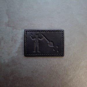 Op de afbeelding: Zwarte leren patch met een gestikte rand. De patch heeft een gestileerde afbeelding van een skelet dat een zwaard en een hart vasthoudt.