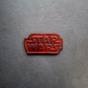 Op de afbeelding: Een bruin leren patch met de woorden "STAR WARS" in reliëf in een donkerdere bruine kleur.