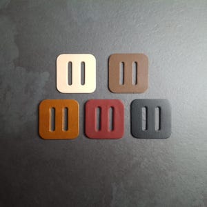 Puede incluir: Cinco hebillas cuadradas de cuero en varios colores: crema, marrón, beige, burdeos y negro. Cada hebilla tiene dos ranuras verticales. Las hebillas están dispuestas sobre una superficie gris oscuro.