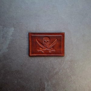Peut inclure: Un patch en cuir marron avec un motif de crâne et d'os croisés. Le crâne est blanc et les os croisés sont noirs.