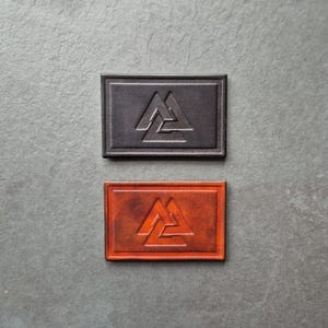 以下が含まれることがあります： 3つの三角形が重なり合ったデザインが浮き彫りになった革のパッチが2つ。パッチは長方形で、縁取りがあります。1つのパッチは黒革、もう1つは茶革です。