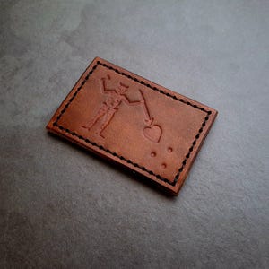 Op de afbeelding: Een bruine leren patch met een gestikte rand. De patch heeft een reliëfbeeld van een stokfiguur dat een speer vasthoudt en naar een hart wijst. Drie stippen bevinden zich onder het hart.