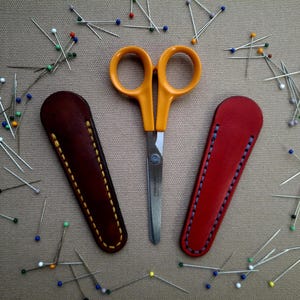 Puede incluir: Unas tijeras con mango naranja y hojas plateadas, flanqueadas por dos fundas de cuero, una marrón y otra roja. Alfileres de costura con cabezas de colores están esparcidos alrededor de las tijeras y las fundas, sugiriendo un tema de costura o artesanía.