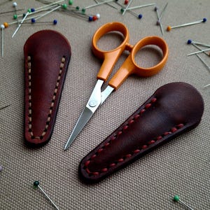 May include: Orange-handled scissors with silver blades and two brown leather sheaths with red stitching. Sewing pins with colorful heads are scattered around the scissors and sheaths. The items are on a tan surface.