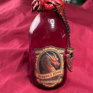 Puede incluir: Una botella de vidrio burdeos con un sello de cera roja y una cadena decorativa. La etiqueta dice "Dragon's Blood" con una ilustración de dragón. La botella tiene un dije decorativo de punta de flecha.