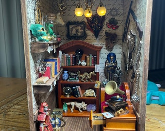 May include: A miniature dollhouse room with a wooden floor, a bookshelf filled with books, a small table with a record player, and a samurai figurine standing in the corner. The room is decorated with various trinkets and a chandelier.
