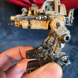 Könnte beinhalten: Ein detailliertes, handbemaltes Spielzeugroboter im Steampunk-Stil mit einem beige-braunen Farbschema. Der Roboter hat einen großen, detaillierten Kopf mit einem großen, runden Fenster und einen kleinen, detaillierten Körper mit einem großen, runden Fenster. Der Roboter hat eine große, detaillierte Waffe auf dem Kopf. Der Roboter steht auf einem Paar großer, detaillierter Ketten.